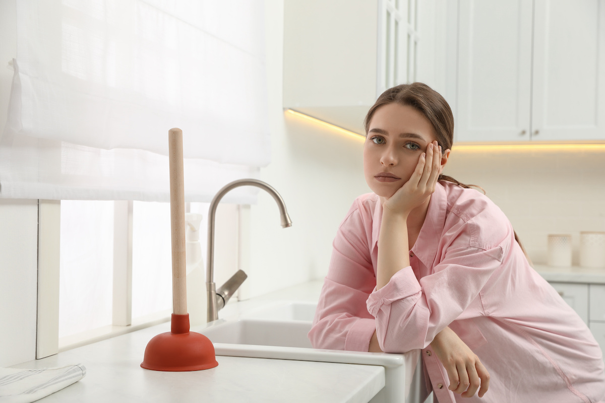 Unhappy young woman with plunger near clogged sink in kitchen
